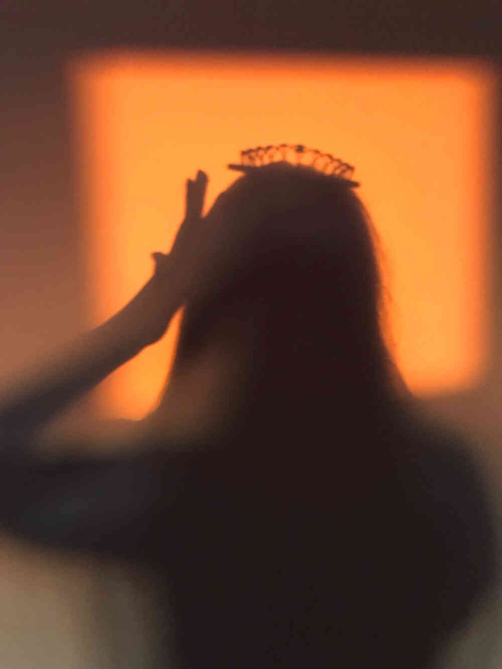 Silhoutte from behind, against bright orange backdrop, of long-haired woman touching her head and with a crown on her head.